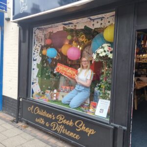 External window of Wonderland Bookshop. Author LD Lapinski is sitting inside the window holding a sign saying The Strangeworlds Travel Agency